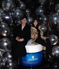 Two people seated on a large skincare product container surrounded by reflective silver balloons at an event space.
