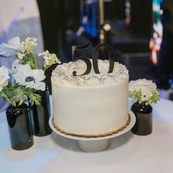 White frosted cake with '50' topper, displayed on a stand on a table alongside small black vases holding white flowers.